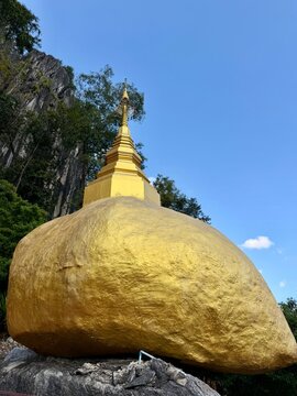 A sacred golden painted boulder topped with a small golden pagoda set against a dramatic limestone cliff and blue sky at Wat Tham Chetawan in Nan province, Thailand.