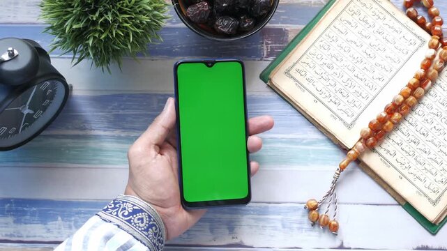 A man's hand holds a smartphone with a green screen above a table with dates, an islamic prayer book, and rosary beads