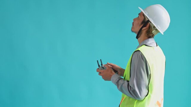 Side view of engineer in hardhat operating drone controller and looking upward on cyan background