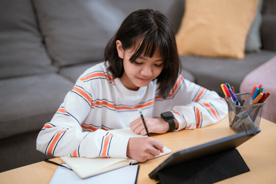 Young girl studying and taking notes while using a tablet at home in a cozy living room