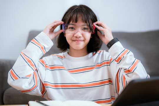 Girl wearing glasses studies at a desk with a tablet and open book in a cozy indoor setting during the afternoon