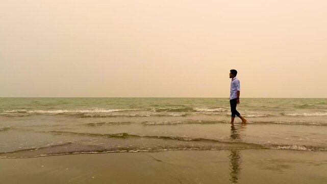 Loneliness concept, solitary man walking along the shoreline at Cox's Bazar sea beach under a hazy sky, gentle ocean waves crashing on the sand, Bangladesh, wide shot, low angle shot.