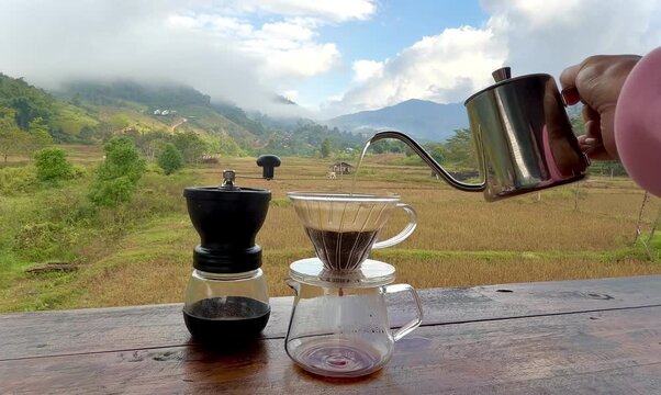 Pouring hot water from a kettle into a glass dripper to brew pour-over coffee with a mountain and valley backdrop in Sapan, Thailand.