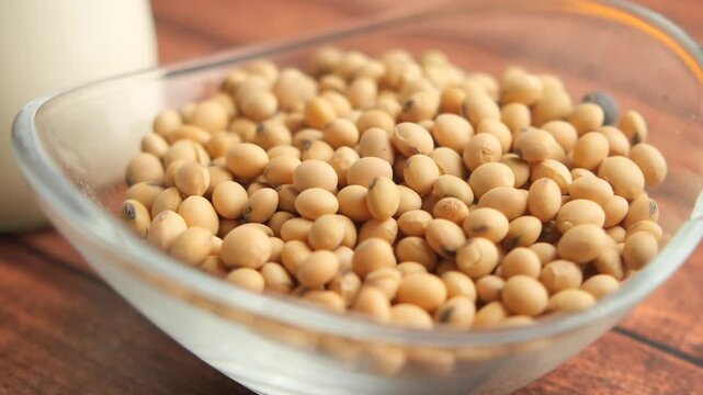 Fresh organic soybeans falling into glass bowl on wooden table surface with soy milk in background