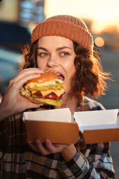 woman burger eating outdoors on street with takeout and beanie, candid lifestyle and authenticity captured at golden hour glow, emotional storytelling of mindful living and cozy urban food moment