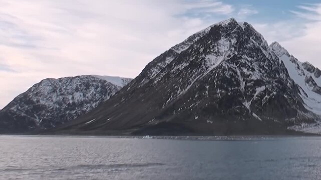Majestic snow-covered mountains rise from the tranquil Arctic Ocean. This epic landscape, seen during an extreme expedition, showcases nature's grand design on a cloudy Arctic day.
