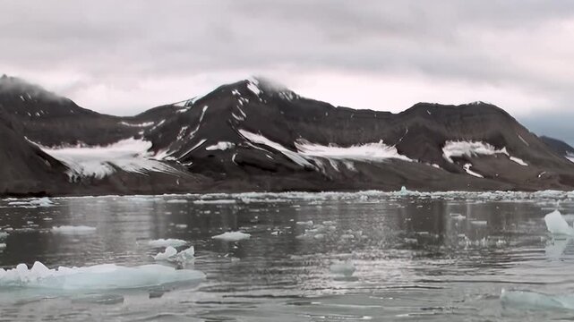 Breathtaking expanse of Arctic Ocean unfurls, dotted with ancient ice floes. Majestic snow-capped mountains loom under overcast sky, reflecting their stark beauty on frigid, pristine waters.