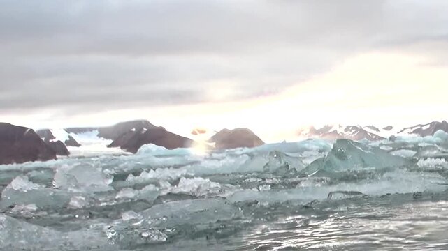 A vessel glides through the shimmering Arctic Ocean, pushing aside fragmented ice. Towering mountains pierce the cloudy sky as golden light illuminates this polar expedition.