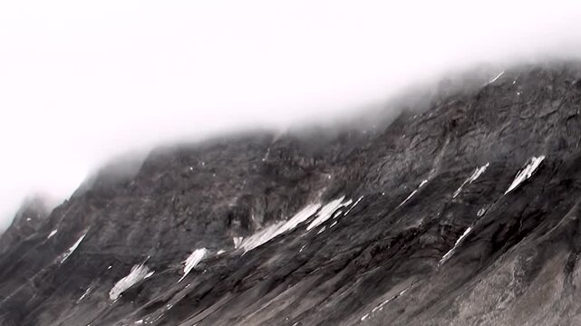 Rugged Arctic mountains emerge from swirling ocean mists. Patches of snow cling to dark, ancient rock, painting a challenging environment for extreme polar travel and exploration.