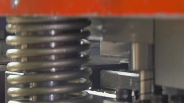 Silver metal spring undergoes rhythmic compression cycles within an active mechanical manufacturing plant during daytime hours to facilitate precise equipment performance and constant machine pressure