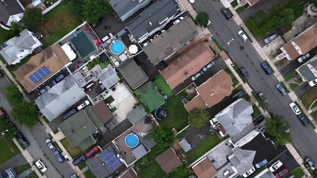 Round swimming pool sits behind dense rooftops in residential neighborhood of Belleville New Jersey, parked cars line narrow street between backyards with solar panels. Aerial drone top down shot