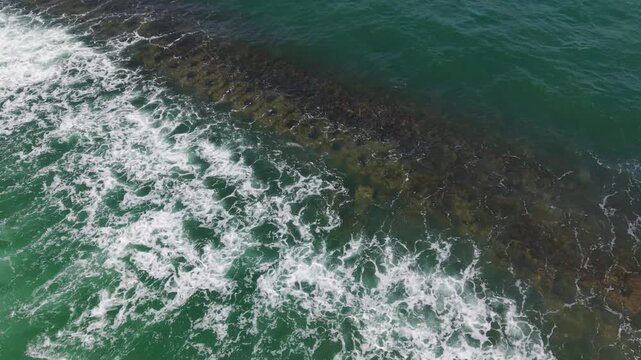 Aerial View Of Rocky Reef, Turquoise Water Crashing Against Submerged Breakwater, Frothy White Foam Tracing Repeating Stone Pattern, Drone Perspective Revealing Coastal Motion, Shallow