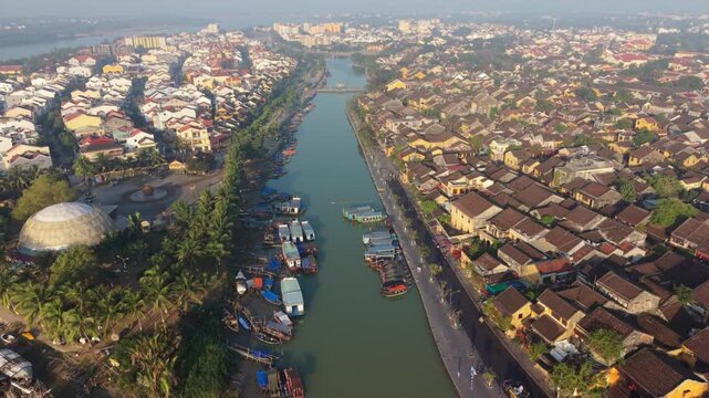 Heritage Quarter Canal In Hoi An Midday, Narrow Waterway Framed By Tiled Roofs And Traditional Houses, Colorful Boats Moored Along Banks And Bustling Streets Visible From Above