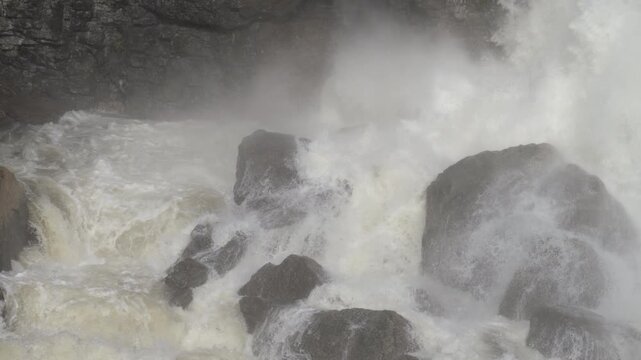 Rushing spring water slams violently against massive dark boulders at the base of Inglis Falls. Heavy mist rises from the turbulent plunge pool, creating a damp atmosphere.