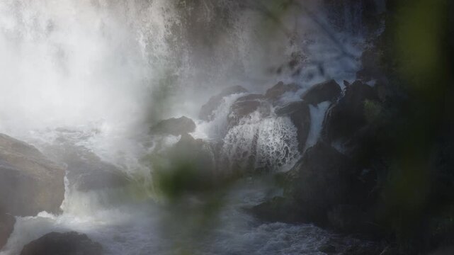 Heavy mist and water spray envelope dark rocky cliffs at Inglis Falls in Owen Sound, Ontario. Out-of-focus green foliage in the foreground frames the powerful cascade.