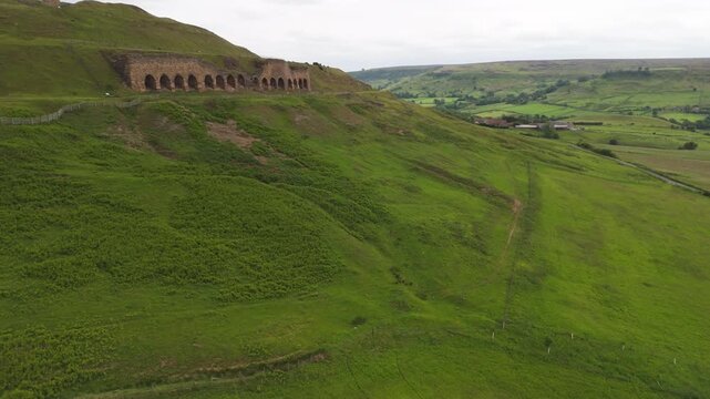 Aerial drone view flying around the abandoned historic stone ironstone kilns in Rosedale Abbey, set against overcast rolling Yorkshire hills.