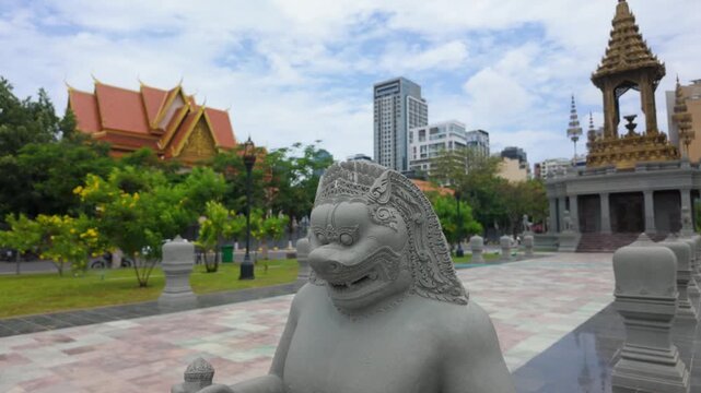 Guardian lion statue at the Norodom Sihanouk Memorial park in Phnom Penh, Cambodia, with the memorial pavilion, Royal Palace roof and modern city towers.
