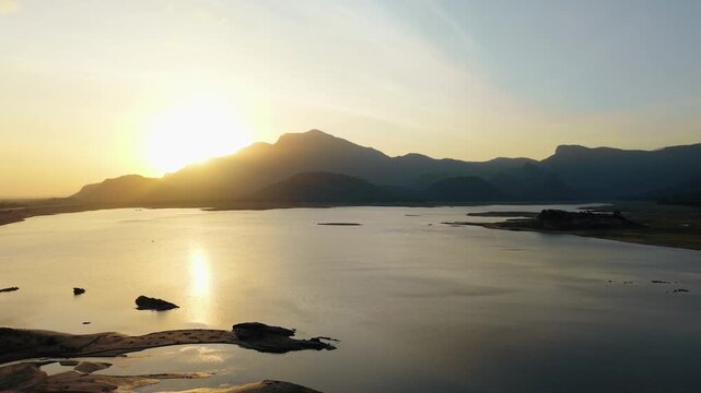 Cinematic aerial drone shot of a calm reservoir at sunrise in South India, golden light glowing behind silhouetted mountains, rocky islets and sandbars scattered across still reflective water.