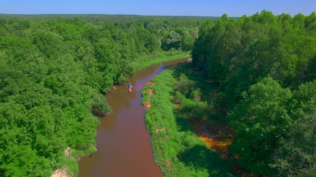 Friends kayaking features colorful inflatable boats following one another around a shaded bend as an elevated drone tracks their movement between dense trees bright grass and calm brown water