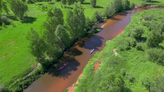 Friends tandem kayaking on an amber river features two inflatable boats traveling downstream between banks and scattered trees while a high oblique drone tracks their parallel course in summer