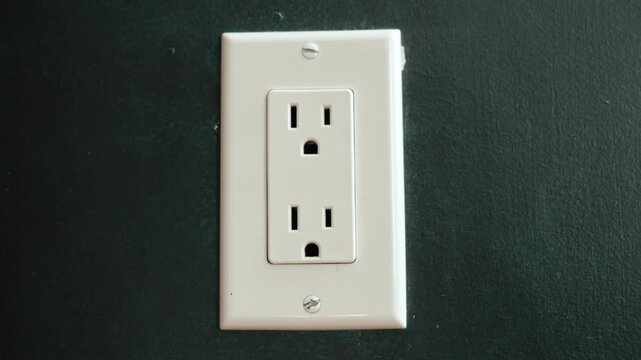 A close up view of an electrical outlet on a dark wall as a hand plugs in an electrical cord.
