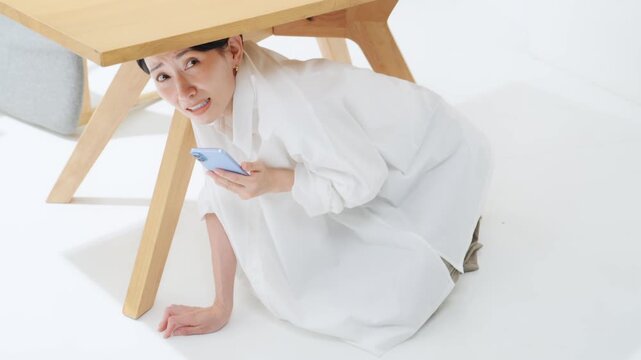 テーブルの下に潜ってスマホを見る女性。地震、緊急地震速報、アラート、防災、災害、怖い、怯える