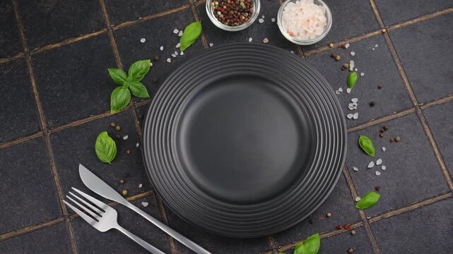 A black plate with fork and knife surrounded by basil leaves and spices on a dark tiled background