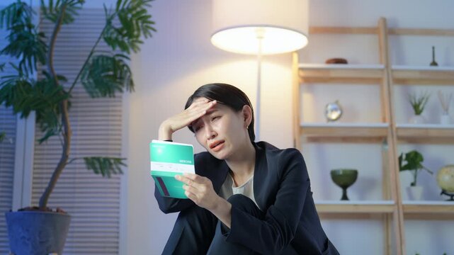 暗い部屋で通帳を見て落ち込むミドル女性。貯金、不安、心配、銀行、家計、投資、資産運用、物価、増税
