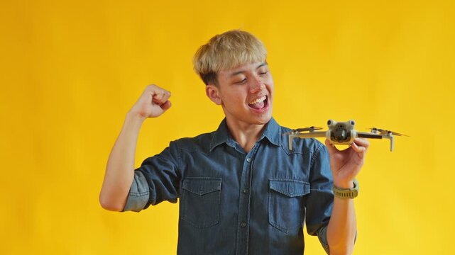 Excited young man holding compact drone and pumping his fist cheerfully against yellow background