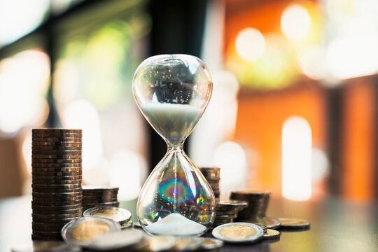 Time is money financial management concept with hourglass standing among stacks of coins on table with soft warm bokeh background for investment planning strategy