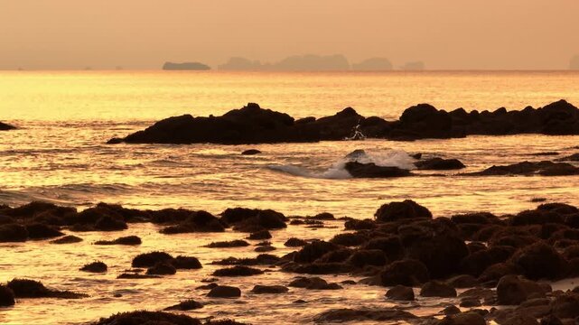 Andaman Sea coastline rocky beach formations and gentle waves reflect the last golden light of the sunset on Koh Lanta, Thailand, creating tranquil nature scene. Islands in the background. Static shot