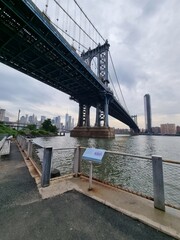 Fototapeta premium Low angle view of the Manhattan Bridge and New York City skyline from Brooklyn waterfront walkway under a cloudy sky