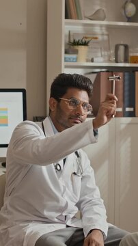 Vertical video of young adult South Asian doctor holding clipboard, then picking up reflex hammer to conduct neurological exam. Clip is ideal for healthcare services and medical training promotions