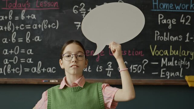 Portrait of Caucasian child holding blank speech bubble cutout in front of blackboard, looking up thoughtfully and then turning to look at camera. Ideal for educational marketing campaigns