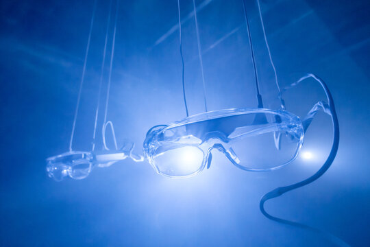 Clear plastic laboratory safety goggles glasses hanging on wires from ceiling in dark blue smoky atmosphere room lit by bright spotlight beam flares, medical hazard scientific research concept.