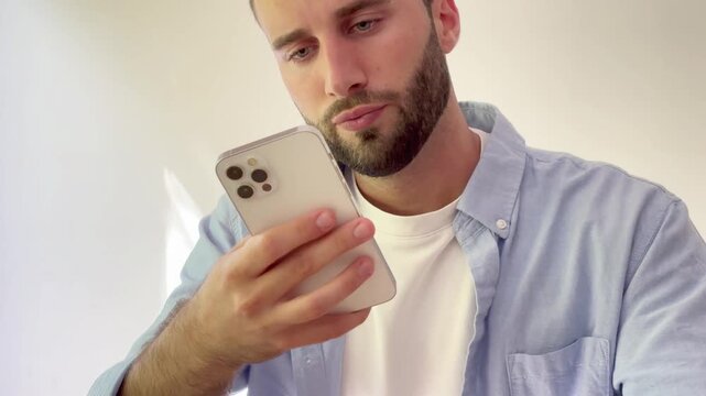 Bored and disappointed young bearded man scrolling on his smartphone, feeling sad while browsing social media feed or waiting for a message on a white background, showing signs of doomscrolling
