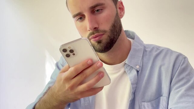 Young handsome man with a beard feeling upset and disappointed while looking at his smartphone screen, reacting to bad news or a negative message isolated on a white background