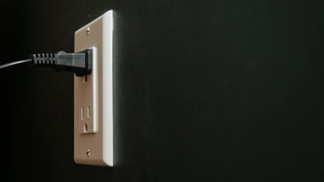 Person inserting and removing black power cord into electrical wall outlet for charging home electronics.