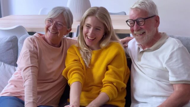 Cheerful multigenerational family with senior parents and an adult daughter sitting together on a sofa, happily waving and smiling during a video call with relatives in their living room