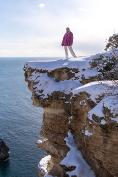 Young woman standing on snowy cliff edge overlooking blue sea during bright winter day