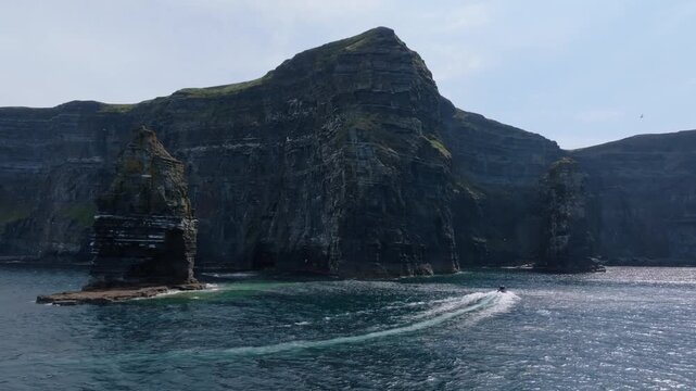 Fast inflatable RIB arcs a white wake around an offshore sea stack beneath sheer Cliffs of Moher on a clear day.