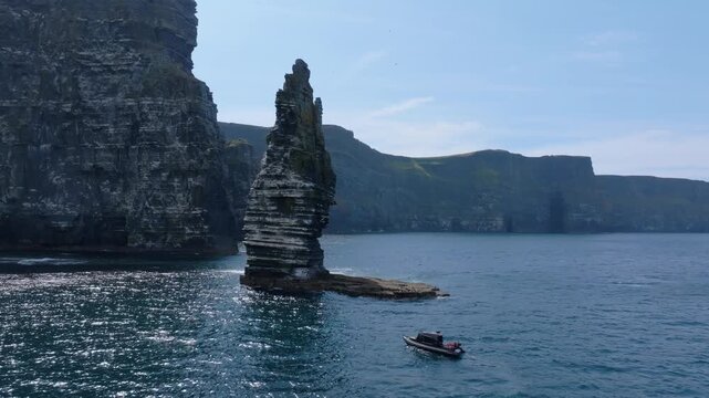 Small RIB tour boat motors past a layered sea spire beneath high grassy cliffs at the Cliffs of Moher, seen from above.