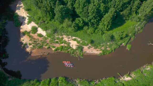 Friends kayaking on a winding forest river places four inflatable kayaks beside a curved sandy point as the drone rises and pulls back to reveal brown water dense woodland and the complete bend