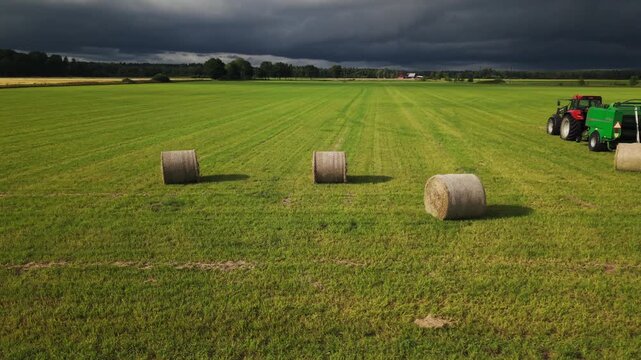 Aerial view of round hay bales and a parked tractor with baling equipment in a green field beneath dark storm clouds, illustrating forage production and weather exposure.