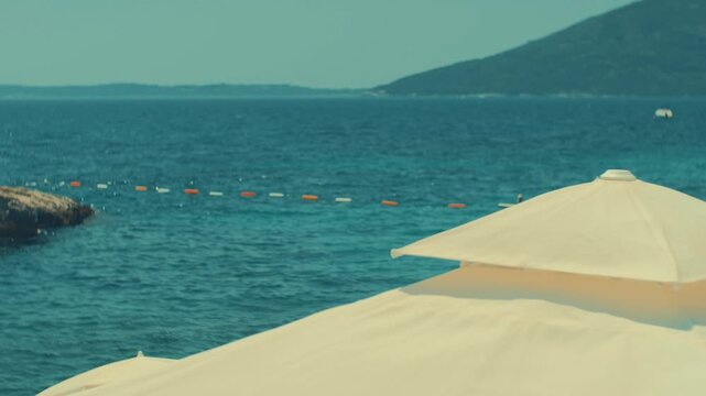 Cinematic vintage footage of a beach sun umbrella in the foreground overlooking the blue sea, rocky coastline, and distant mountains on a sunny summer day.