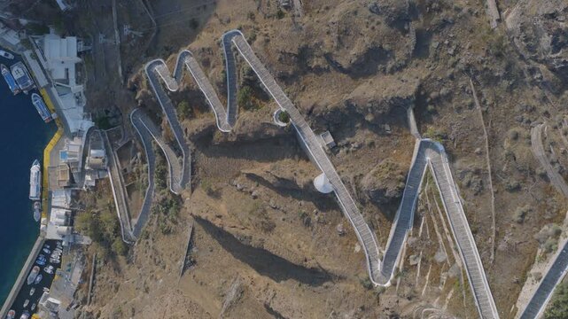 A top-down drone view reveals the famous zigzag switchback path carved into Santorini's caldera cliff, connecting the port to Fira above.