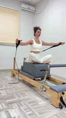 Fototapeta premium Athletic woman training pilates on wooden reformer in bright studio while holding handles for upper body resistance during active workout