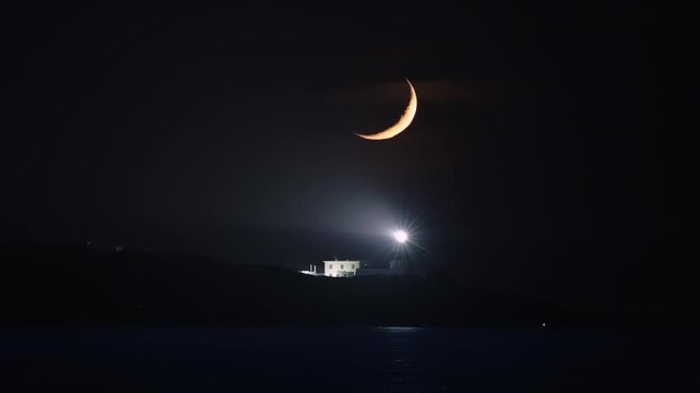 A glowing crescent moon hangs in the dark night sky above the flashing light of Fugui Cape Lighthouse in Shimen, New Taipei City, Taiwan.