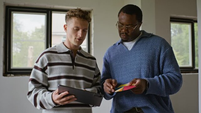 Young adult Caucasian anf African American male designers discussing color swatches and using tablet in unfinished room. Footage is perfect for interior design and home renovation marketing