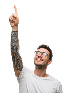 Smiling young caucasian man in a white t-shirt and glasses, detailed sleeve tattoo visible, pointing index finger up on an infinite bright white studio background with copy space. Concept of